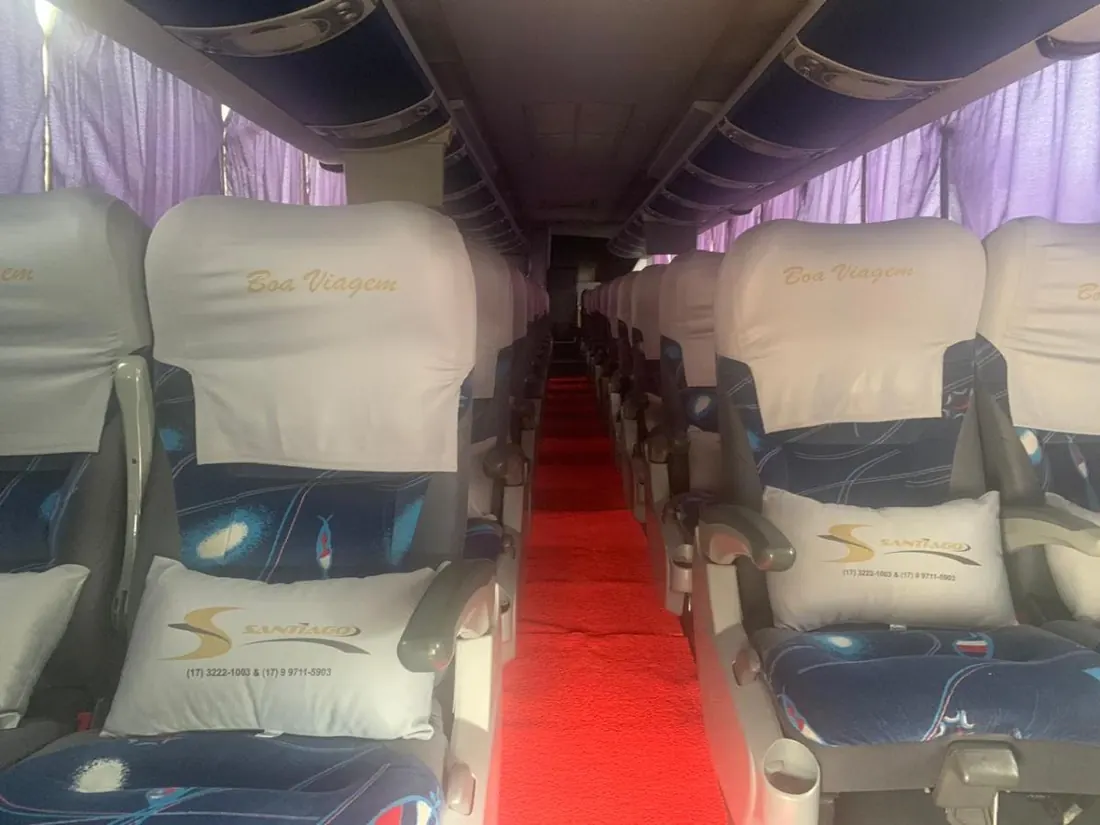 Corredor interno de ônibus da frota, com carpete vermelho, poltronas forradas e almofada bordada com a marca Santiago